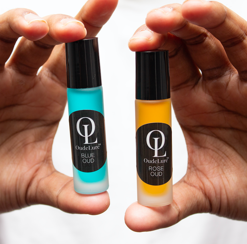 Two small bottles of Oudelure perfume, one blue and one rose-scented, held between fingers against a neutral background.